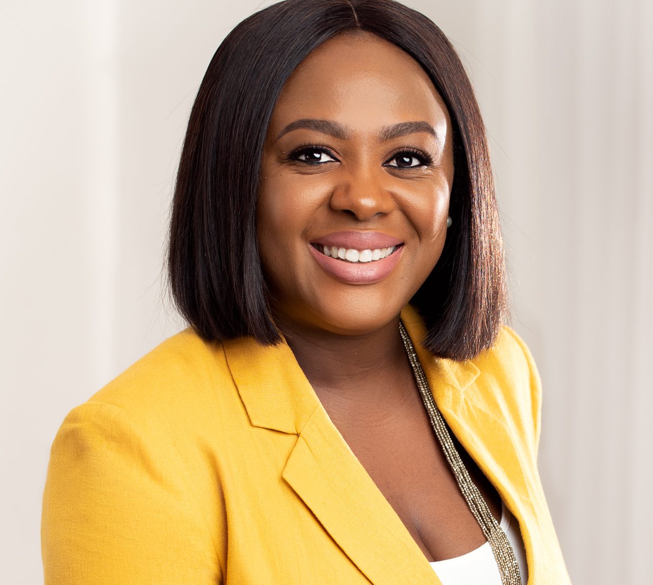 Mojolaoluwa Aderemi-Makinde smiles at the camera. She is wearing a yellow blazer and is a Black woman with a short bob, set against a professional neutral background.